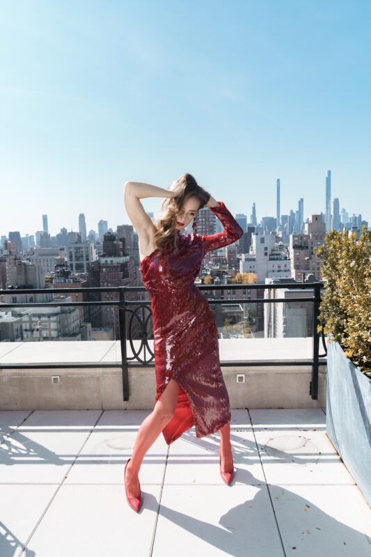 Olga Sorokina in a red dress poses outside with a red rose perfume in her hand