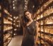A woman in an elegant dress is surrounded by shelves of luxury perfume bottles